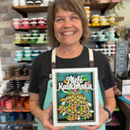 A smiling woman in a black t-shirt and teal apron holds up a framed decorative sign that reads 'Mēle Kaulahāka' above a layered pineapple design. The background shows shelves filled with colorful craft supplies in a workshop or studio.