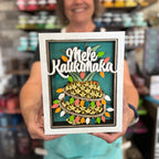 A person holding a framed decorative sign that reads 'Mele Kalikimaka' above a stylized pineapple illustration, with a blurred store background.