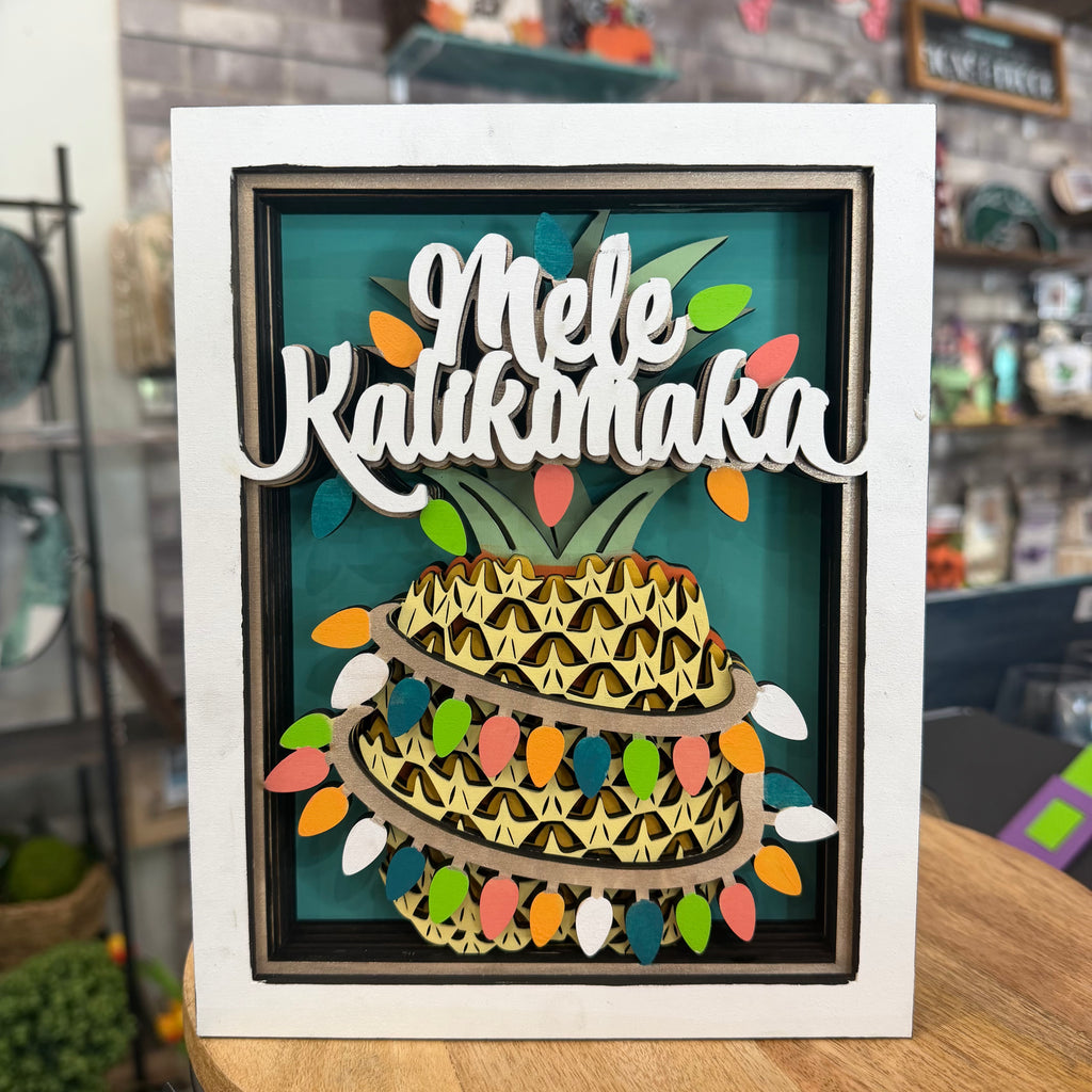 A framed layered wooden art piece featuring a pineapple with the words 'Mele Kalikimaka' in white script across the top, set against a teal background with colorful Christmas lights and ornaments. The piece is displayed on a wooden table in a shop setting.