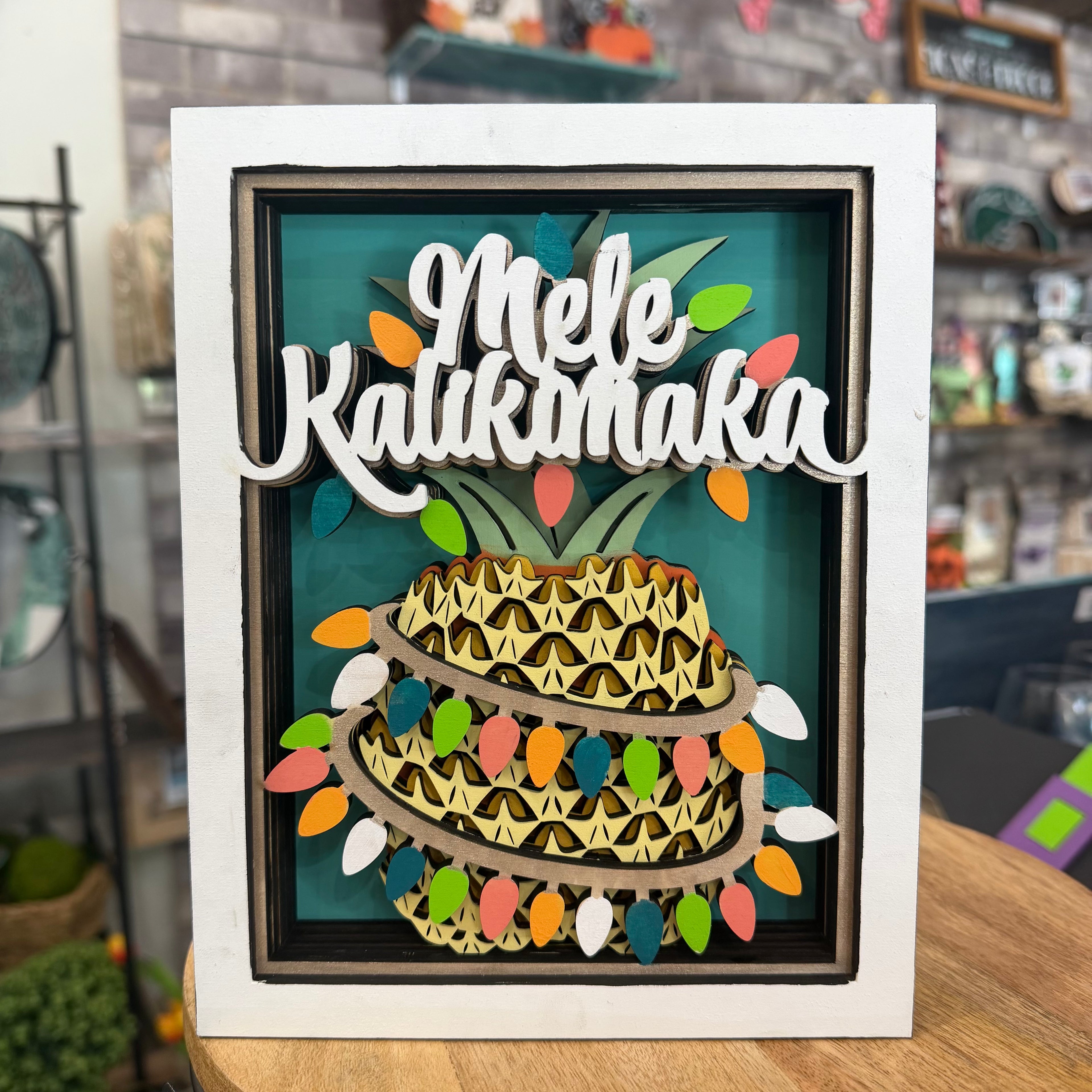 A framed layered wooden art piece featuring a pineapple with the words 'Mele Kalikimaka' in white script across the top, set against a teal background with colorful Christmas lights and ornaments. The piece is displayed on a wooden table in a shop setting.