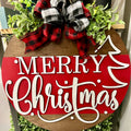 A round door hanger with a Christmas tree design and the text 'Merry Christmas' written in a playful font, decorated with a red and black plaid ribbon on top.