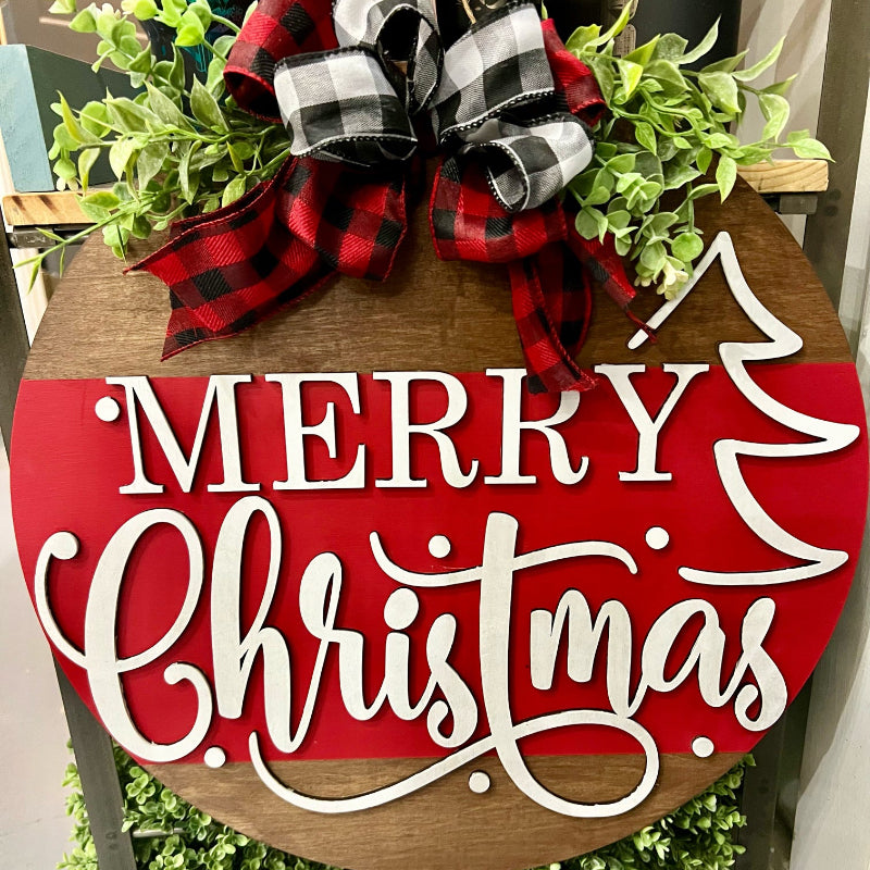 A round door hanger with a Christmas tree design and the text 'Merry Christmas' written in a playful font, decorated with a red and black plaid ribbon on top.