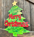 A green and red Christmas tree-shaped paint pour with the text 'Merry Christmas' on it, displayed against a wooden background.