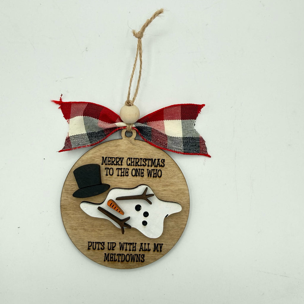 A round wooden Christmas ornament with a red, white, and black plaid ribbon bow at the top. The ornament features a laser-engraved message that reads 'MERRY CHRISTMAS TO THE ONE WHO PUTS UP WITH ALL MY MELTDOWNS' and is decorated with a small 3D snowman figure wearing a black top hat and an orange carrot nose.