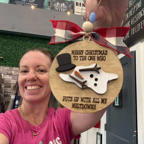 A smiling woman in a pink shirt holds up a round wooden Christmas ornament with a red plaid ribbon. The ornament features a snowman design with a black top hat, orange carrot nose, and black engraved text that reads 'MERRY CHRISTMAS TO THE ONE WHO PUTS UP WITH ALL MY MELTDOWNS'.