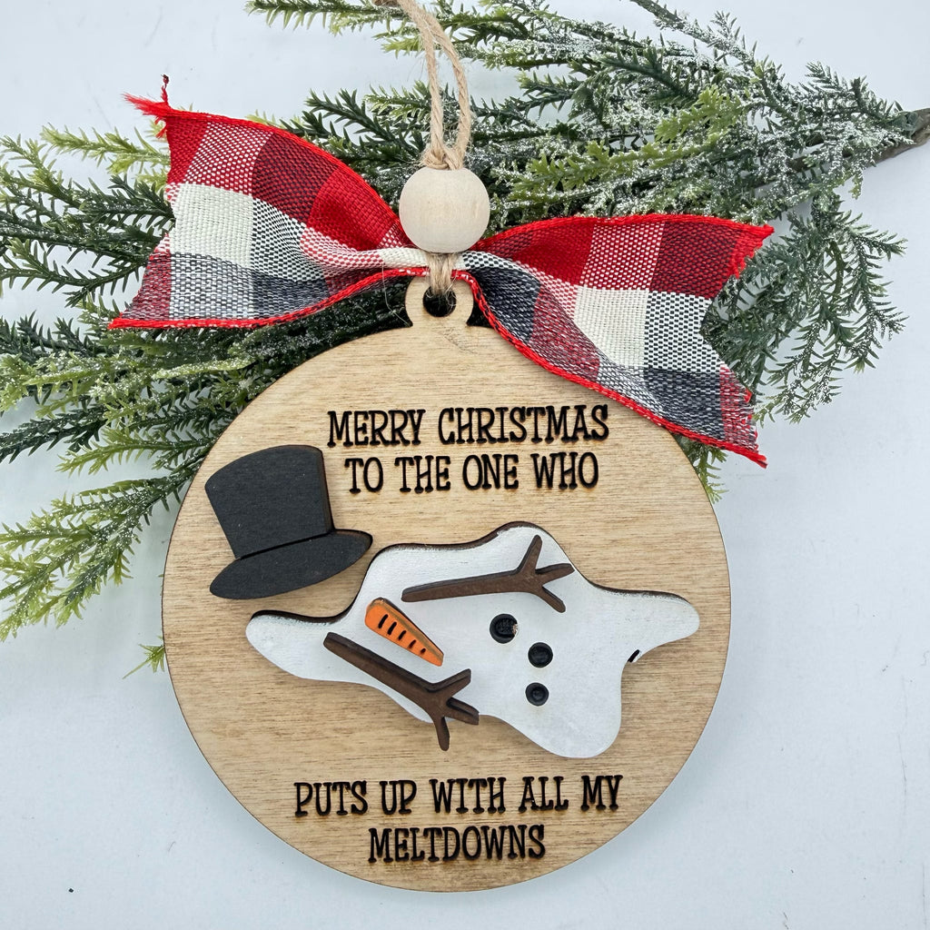 A round wooden Christmas ornament with a laser-engraved humorous message about snowman antics, decorated with a red, white, and black plaid ribbon bow and green pine branches in the background.