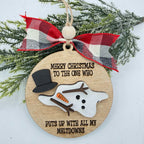 A round wooden Christmas ornament with a laser-engraved humorous message about snowman antics, decorated with a red, white, and black plaid ribbon bow and green pine branches in the background.