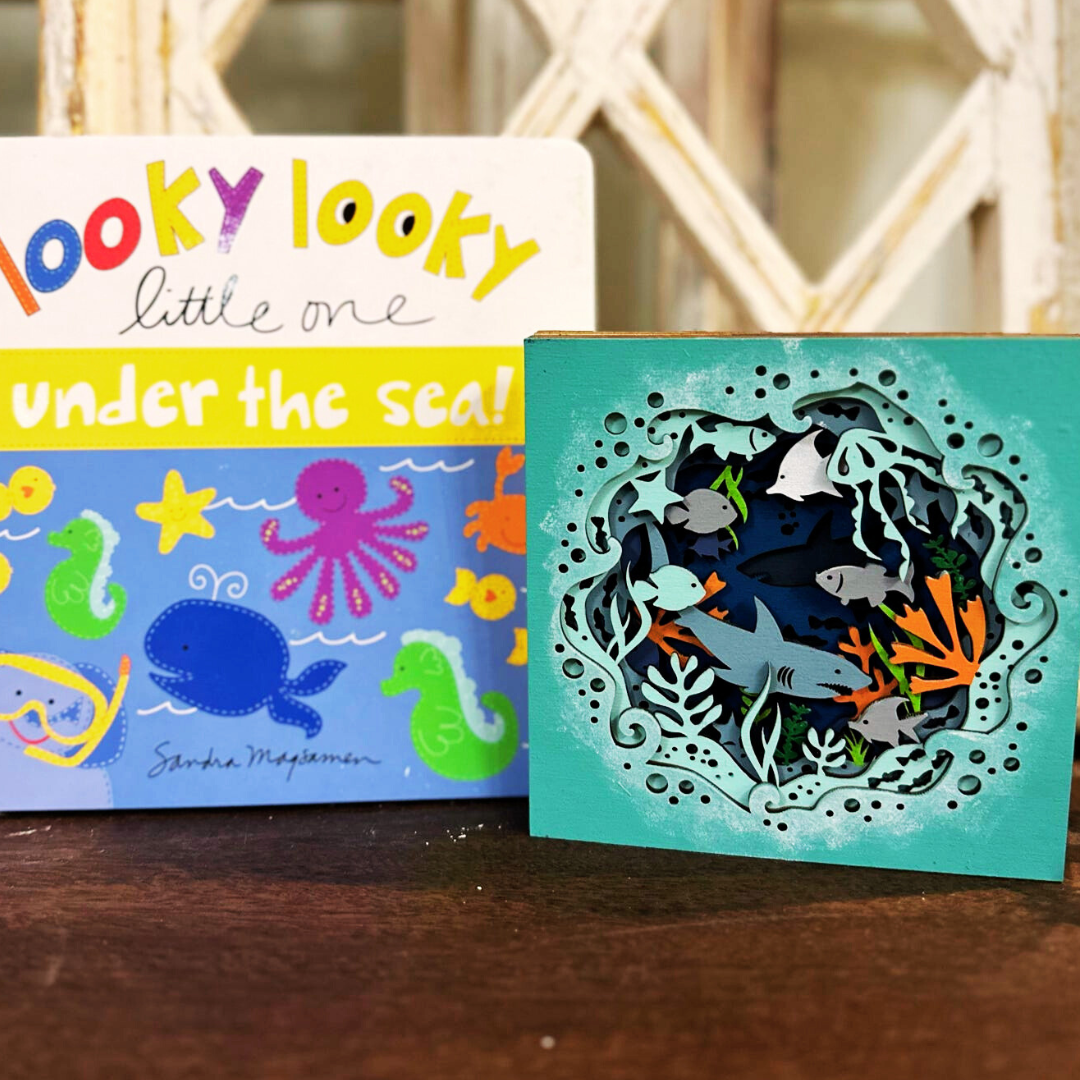 A craft kit depicting an ocean theme with colorful cut-outs of sea creatures, placed next to a colorful book with text and illustrations on the cover.