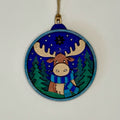Decorative wooden ornament with a moose wearing a blue scarf against a blue sky with stars and green trees.