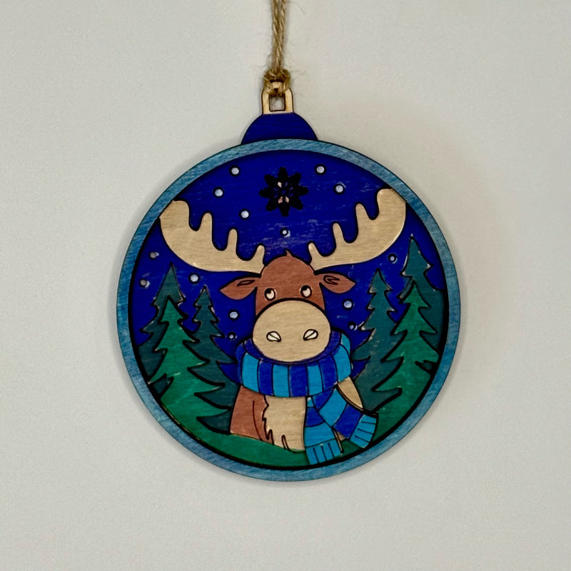 Decorative wooden ornament with a moose wearing a blue scarf against a blue sky with stars and green trees.
