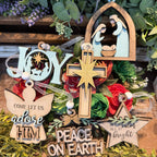 A collection of wooden and metal Christmas ornaments styled with green foliage and red roses on a rustic wooden surface. The ornaments include a wooden nativity scene, a wooden cross with a gold star, a wooden angel ornament, a wooden star, and a light blue 'JOY' letter ornament.