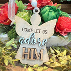 A wooden angel-shaped Christmas ornament with the words 'COME LET US adore HIM' in dark brown lettering, displayed in front of a floral arrangement with red and green roses.