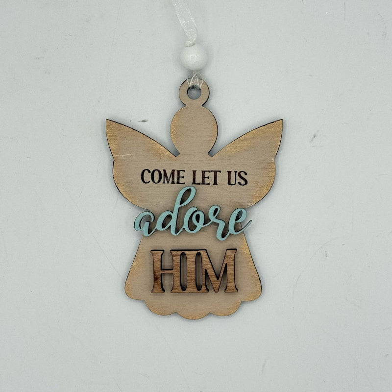 A wooden angel-shaped Christmas ornament with the words 'COME LET US adore HIM' printed on it, hanging against a plain light gray background.