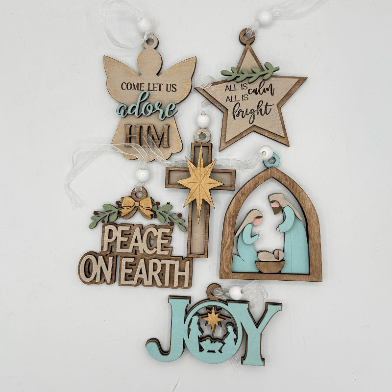 A set of handcrafted wooden Christmas ornaments hanging from white strings against a plain white background. The ornaments include a wooden angel with the text 'COME LET US adore HIM', a star with 'ALL IS color ALL IS bright', a cross, a nativity scene in an arched frame, a 'PEACE ON EARTH' sign, and a 'JOY' ornament with a star cutout.