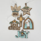 A set of handcrafted wooden Christmas ornaments hanging from white strings against a plain white background. The ornaments include a wooden angel with the text 'COME LET US adore HIM', a star with 'ALL IS color ALL IS bright', a cross, a nativity scene in an arched frame, a 'PEACE ON EARTH' sign, and a 'JOY' ornament with a star cutout.