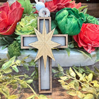 A wooden Christmas cross ornament with a natural wood star in the center, decorated with a small white and blue bead at the top, displayed in a floral arrangement.