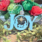 A light blue wooden hanging ornament spelling JOY, with a nativity scene cutout in the center and a clear hanging loop, displayed among faux red roses and green foliage.