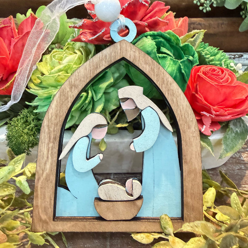 A wooden nativity scene ornament featuring Mary, Joseph, and baby Jesus in a arched frame, displayed in front of a decorative floral arrangement with red roses and green leaves.