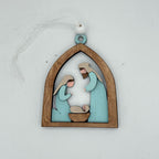 A wooden Christmas ornament depicting a nativity scene with Mary, Joseph, and baby Jesus in a arched frame, hanging by a white ribbon on a light gray background.