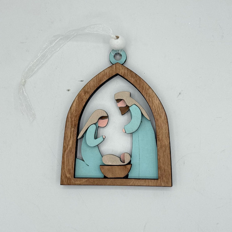 A wooden Christmas ornament depicting a nativity scene with Mary, Joseph, and baby Jesus in a arched frame, hanging by a white ribbon on a light gray background.