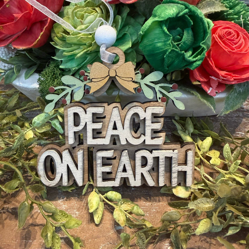 A rustic wooden sign reading 'PEACE ON EARTH' in raised letters, decorated with a small bow, green leaves, and red berries, displayed in front of a floral arrangement with red and green faux roses.