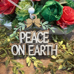A rustic wooden sign reading 'PEACE ON EARTH' in raised letters, decorated with a small bow, green leaves, and red berries, displayed in front of a floral arrangement with red and green faux roses.