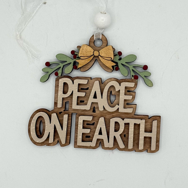 A wooden hanging ornament with the words “PEACE ON EARTH” in raised natural wood letters, decorated with green holly leaves, small red berries, and a gold bow at the top, suspended by a white ribbon and white bead.