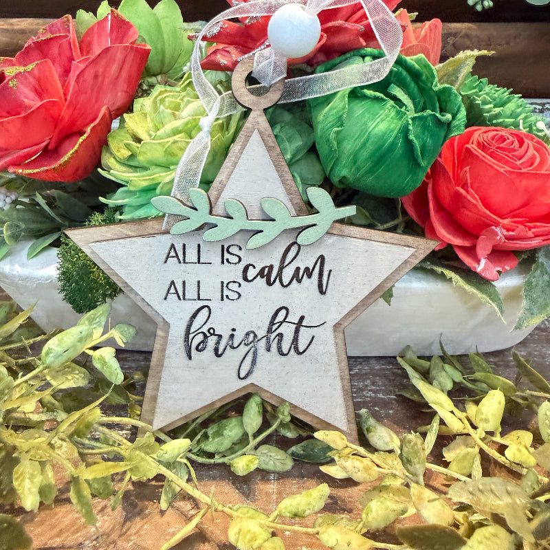 A rustic wooden star-shaped ornament with the phrase 'ALL IS calm bright' in black script, hanging in front of a floral arrangement with red and green flowers and a white bead ribbon.