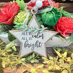 A rustic wooden star-shaped ornament with the phrase 'ALL IS calm bright' in black script, hanging in front of a floral arrangement with red and green flowers and a white bead ribbon.