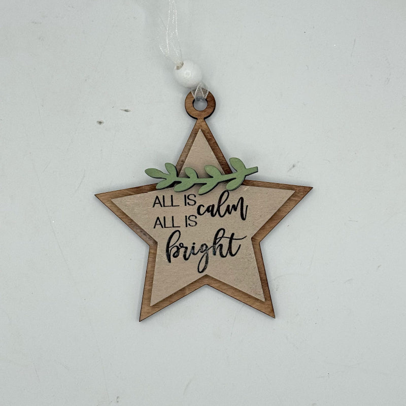 A wooden star-shaped Christmas ornament with a natural wood and light beige center, featuring the text 'ALL IS calm ALL IS bright' in black script, a small green leafy accent, and a white hanging loop at the top, shown against a plain light gray background.