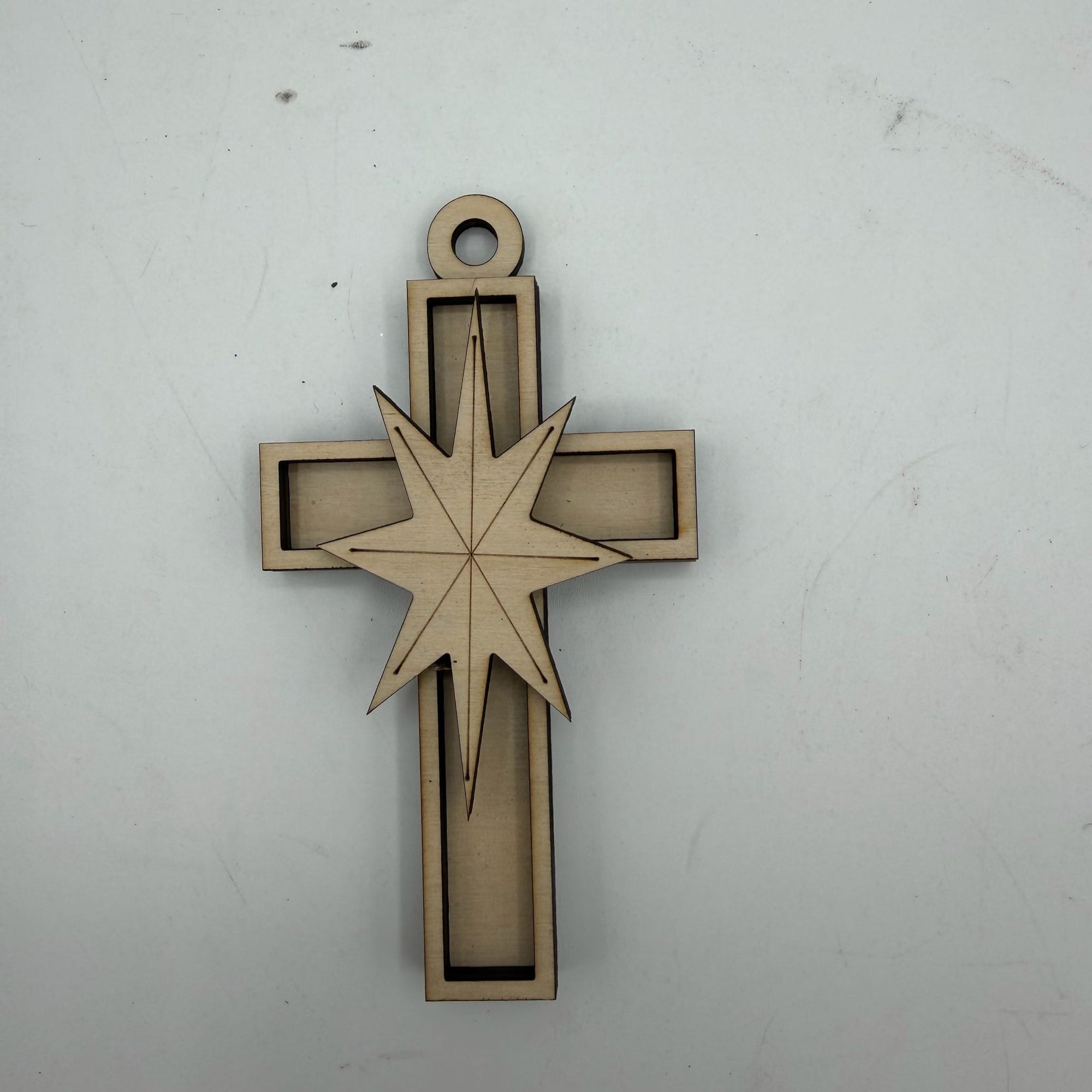 A laser-cut wooden hanging ornament shaped like a cross with a star overlay, shown on a plain light gray background.