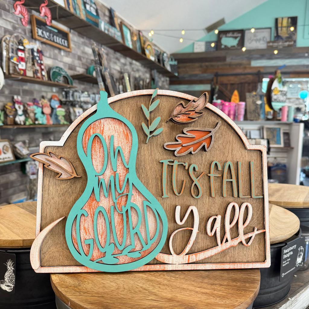 A decorative autumn wooden sign displayed on a wooden table inside a gift shop. The sign features a turquoise gourd silhouette with the words 'ON MY GOURD' over a distressed orange wood-grain background, alongside the phrase 'IT'S FALL Y'all' in matching turquoise and light wood tones, decorated with small orange and brown leaves.