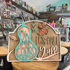 A decorative autumn wooden sign displayed on a wooden table inside a gift shop. The sign features a turquoise gourd silhouette with the words 'ON MY GOURD' over a distressed orange wood-grain background, alongside the phrase 'IT'S FALL Y'all' in matching turquoise and light wood tones, decorated with small orange and brown leaves.