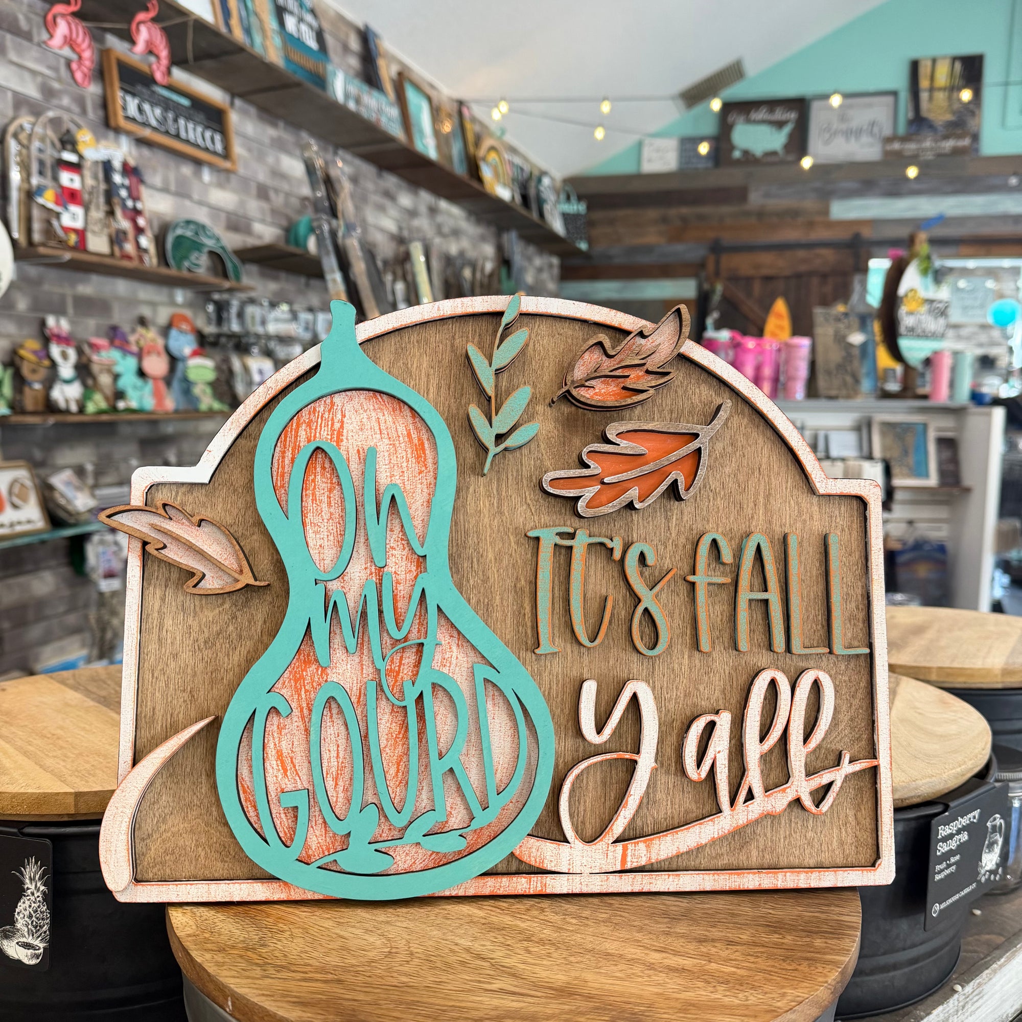 A decorative autumn wooden sign displayed on a wooden table inside a gift shop. The sign features a turquoise gourd silhouette with the words 'ON MY GOURD' over a distressed orange wood-grain background, alongside the phrase 'IT'S FALL Y'all' in matching turquoise and light wood tones, decorated with small orange and brown leaves.