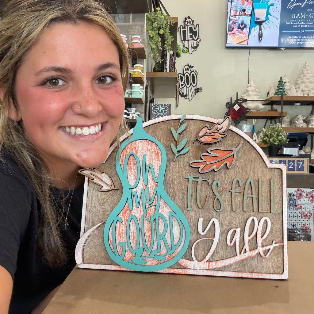 A smiling woman holds a decorative autumn wooden sign that reads 'On My Gourd It's Fall Y'all' in a craft store setting.