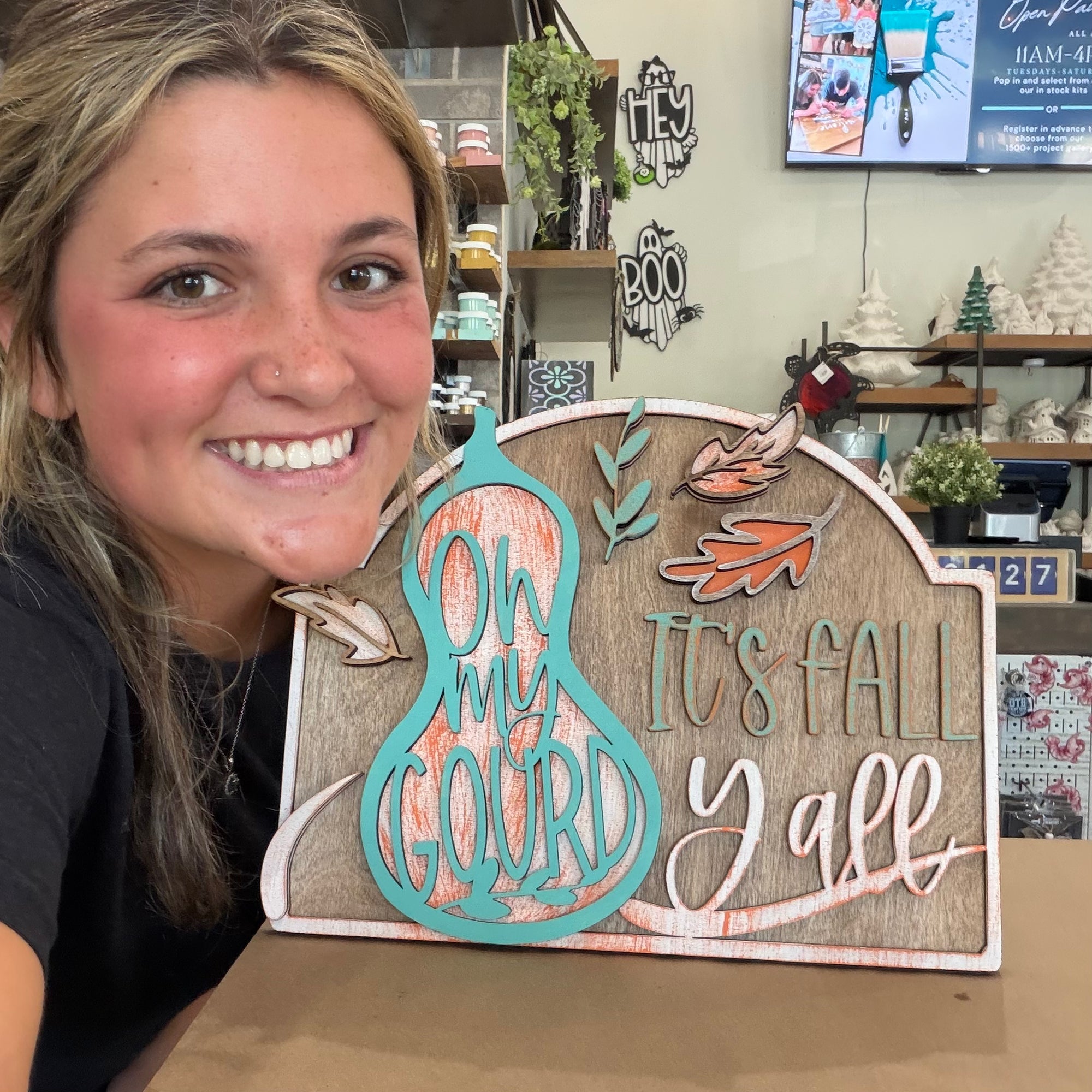A smiling woman holds a decorative autumn wooden sign that reads 'On My Gourd It's Fall Y'all' in a craft store setting.