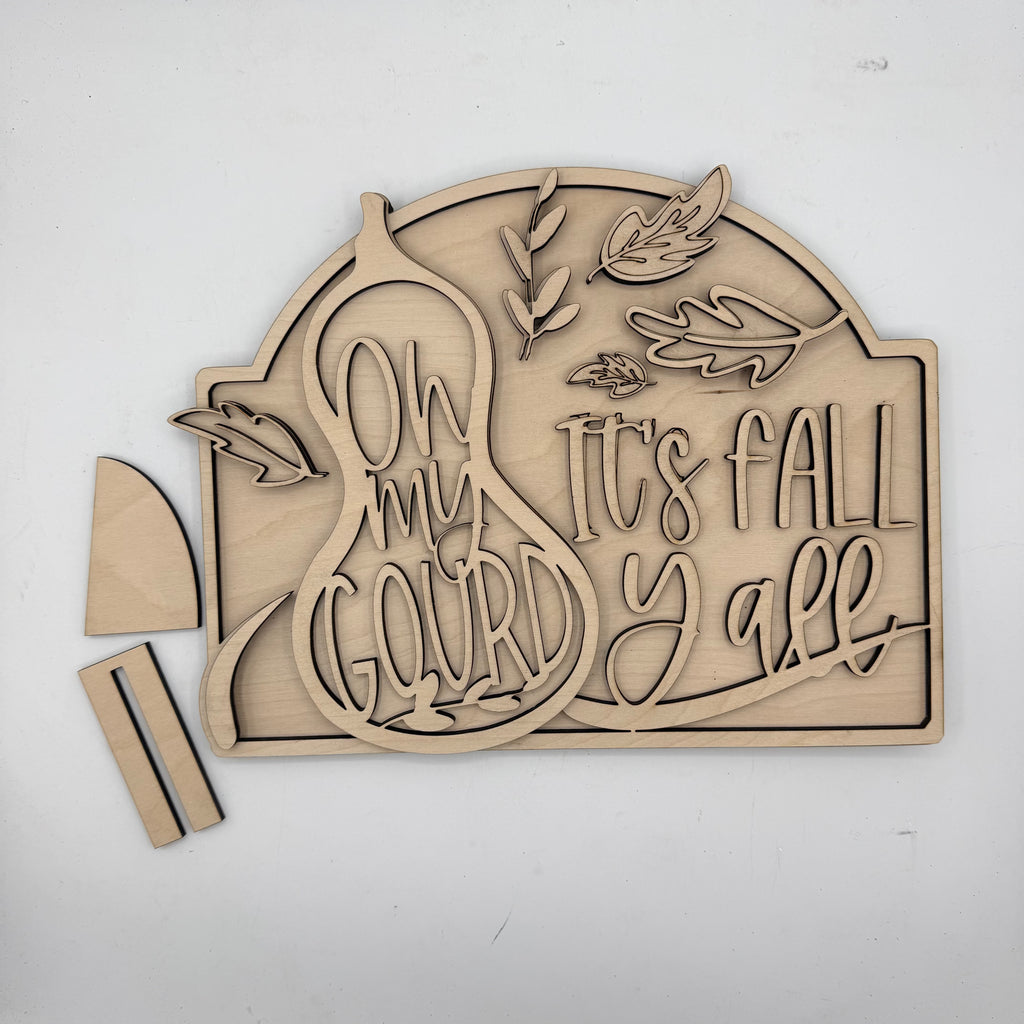 Unfinished wooden laser-cut sign with the phrase 'Oh my Gourd It's Fall Y'all' and autumn leaf designs, shown with two small wooden pieces on a white background.