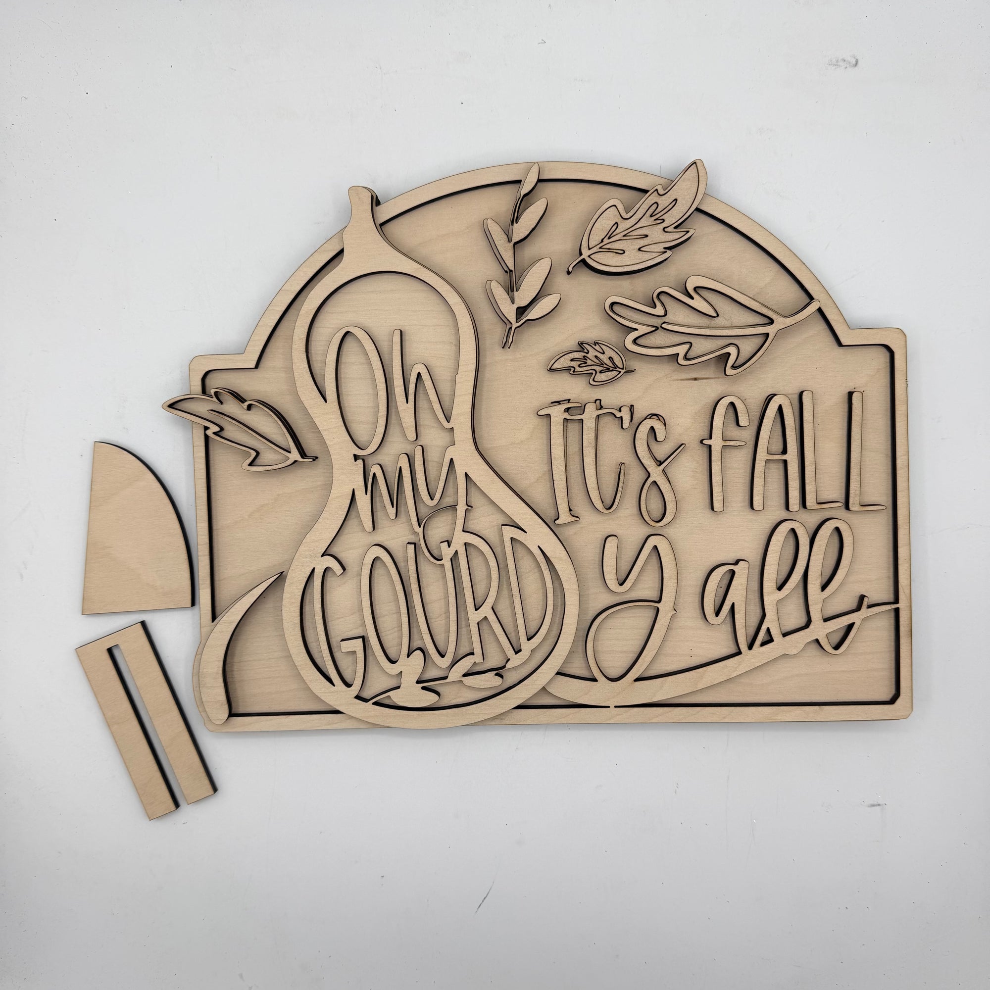 Unfinished wooden laser-cut sign with the phrase 'Oh my Gourd It's Fall Y'all' and autumn leaf designs, shown with two small wooden pieces on a white background.