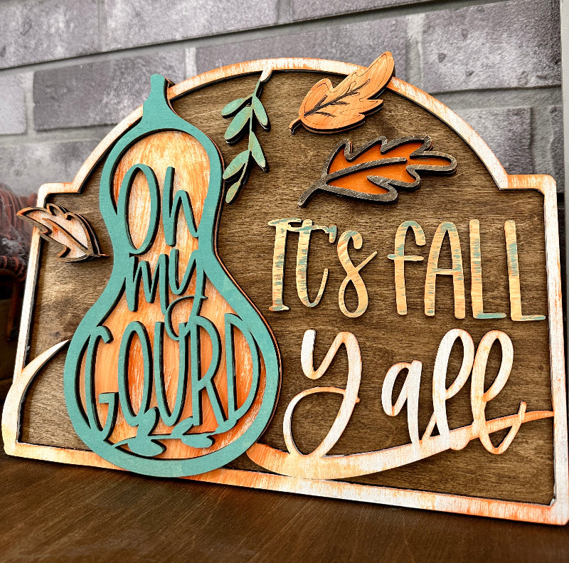 A wooden laser sign with cut-out words and autumn leaf designs, featuring the text 'Oh My Gourd It's Fall Y'all'.