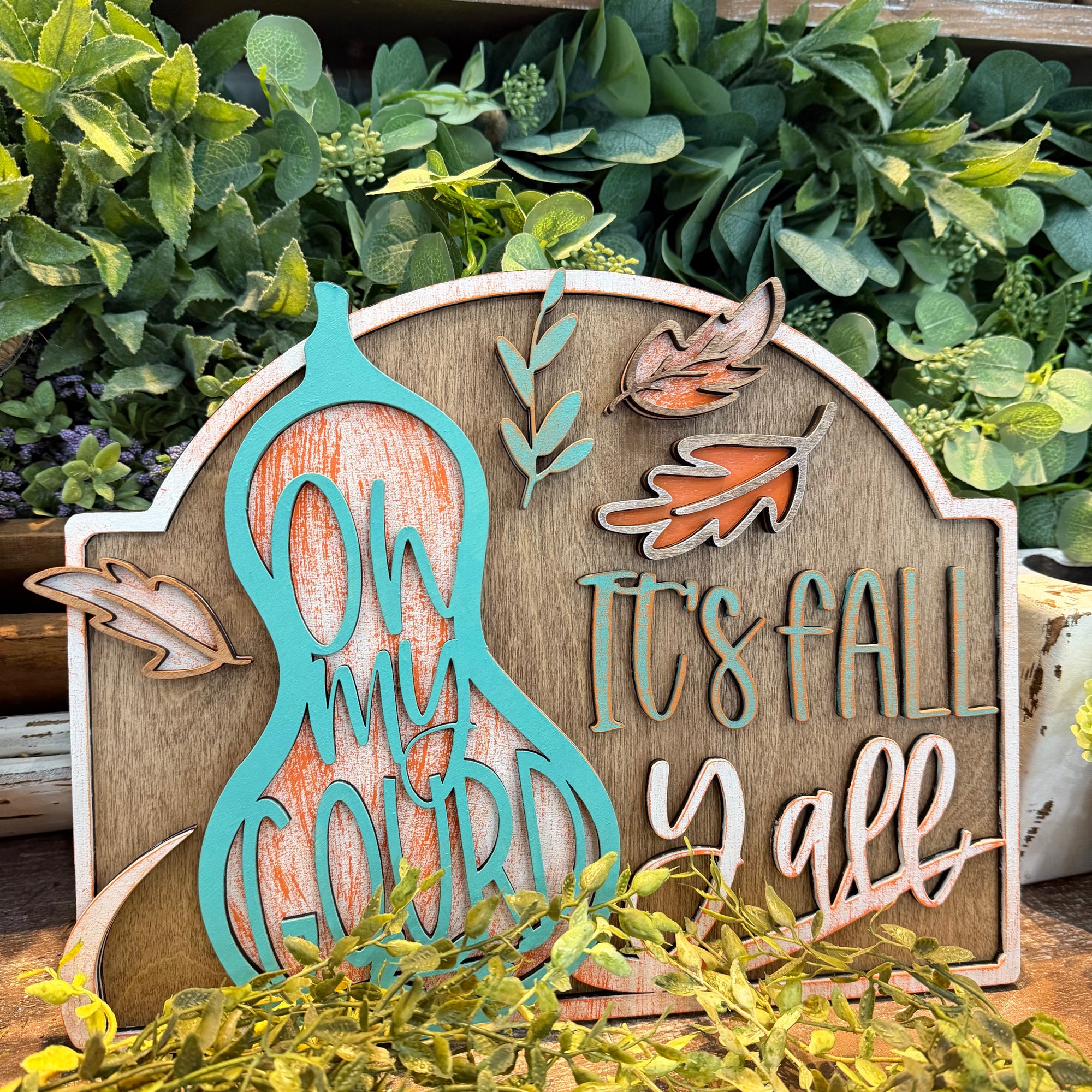 A decorative autumn wooden sign with layered cutout details, featuring a turquoise and orange gourd with the words “Oh my gourds,” fall leaves, and the phrase “It’s Fall Yall.” The sign is styled in front of green foliage.