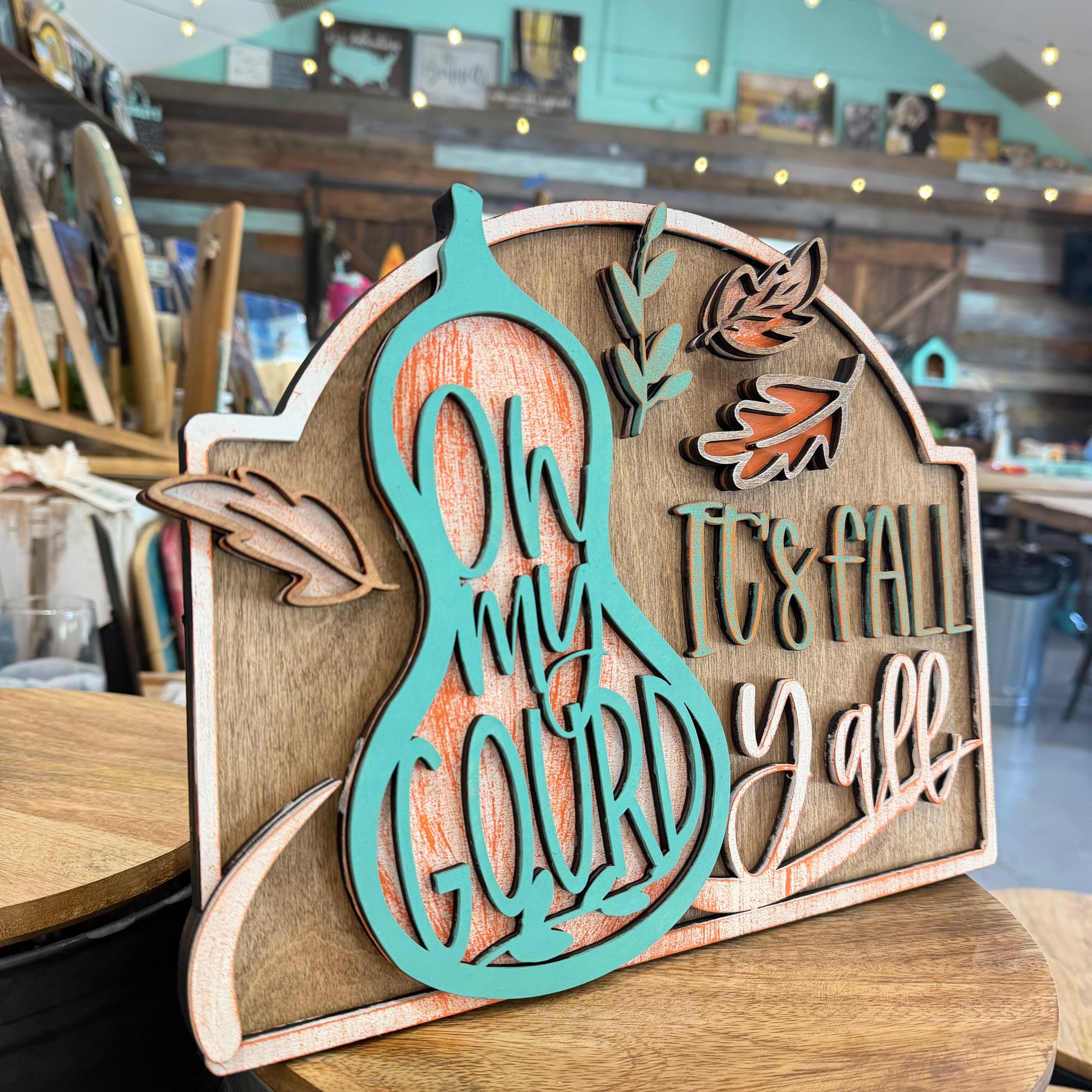 A wooden autumn-themed sign with a turquoise gourd and the words 'Oh my gourd It's fall y'all', displayed on a wooden table in a craft store.