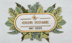 Decorative sign with 'Our Home Est. 2025' text surrounded by green leaves on a white background