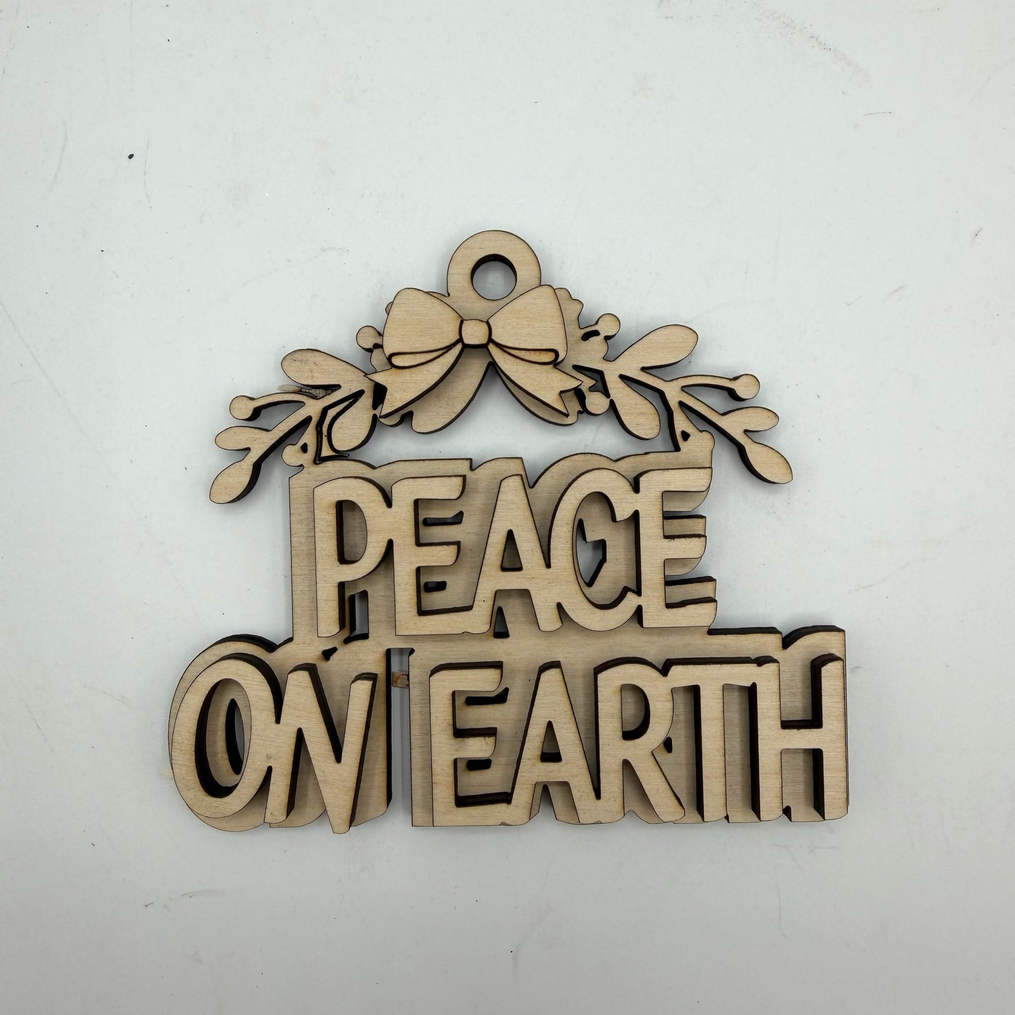 A laser-cut wooden ornament featuring the phrase 'PEACE ON EARTH' in raised letters beneath a bow and holly leaves, shown on a plain off-white background.