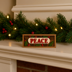 Decorative sign with 'PEACE' on a Christmas mantel with garland and lights.