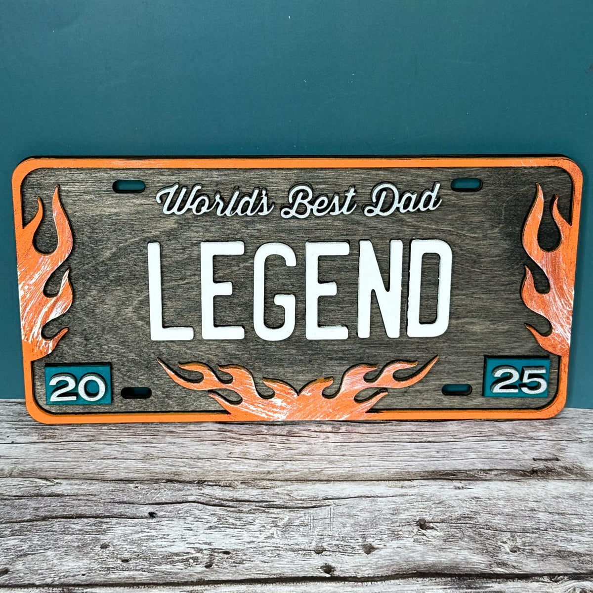 A novelty license plate sign with a wooden background, black and orange text that reads 'WORLD'S BEST DAD LEGEND', and orange flame designs.