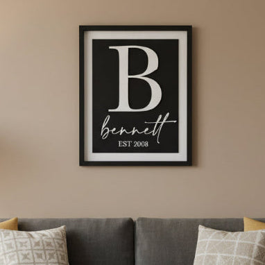 Framed print with a large white letter 'B' and the name 'bennett' with 'EST 2008' below, against a light blue wooden background.