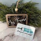Wooden ornament with 'Mrs. Freese' text and a card from Paisley Grace Design on a marble surface with greenery.