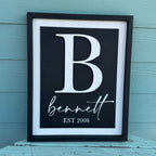 Framed print with a large white letter 'B' and the name 'bennett' with 'EST 2008' below, against a light blue wooden background.