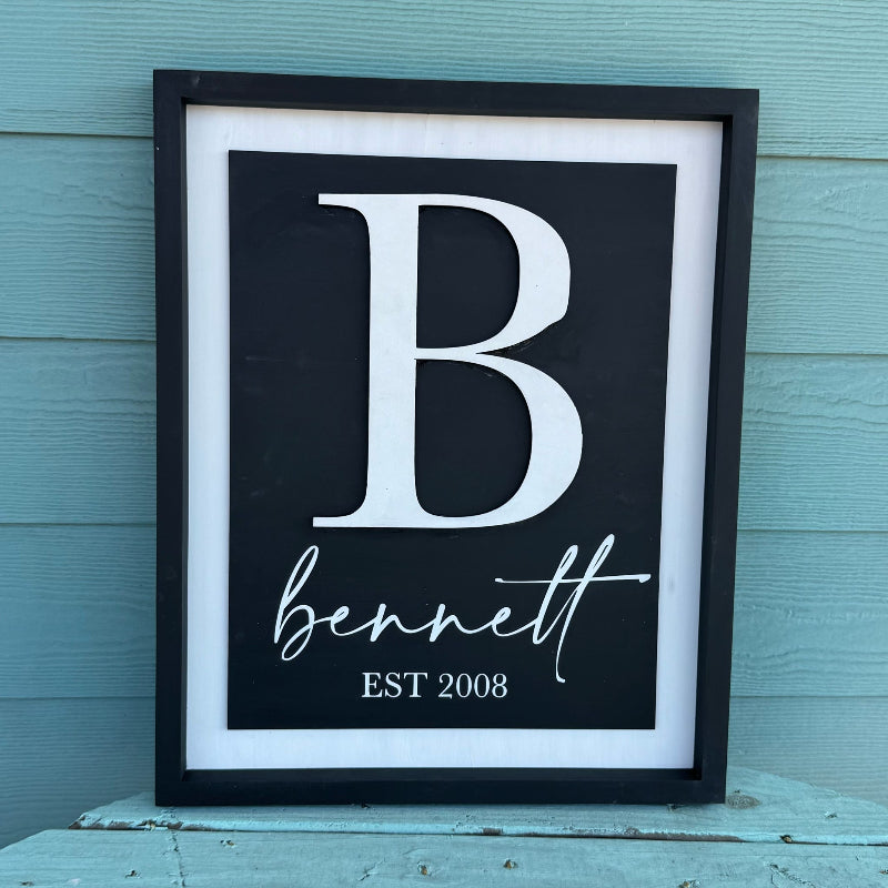 Framed print with a large white letter 'B' and the name 'bennett' with 'EST 2008' below, against a light blue wooden background.