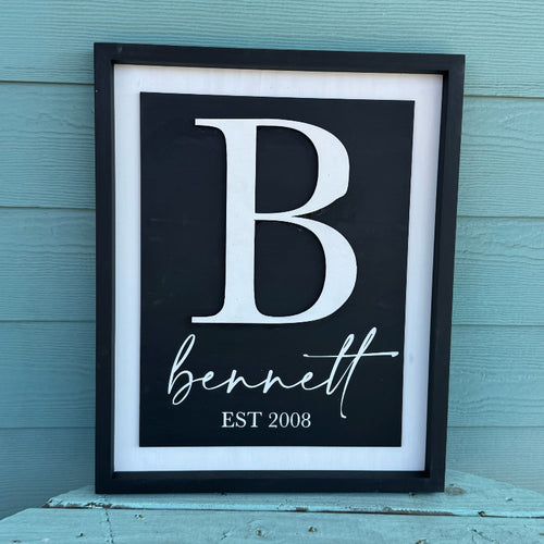 Framed print with a large white letter 'B' and the name 'bennett' with 'EST 2008' below, against a light blue wooden background.